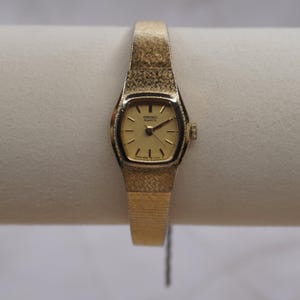 May include: A gold-toned Seiko Quartz watch with a rectangular face and a textured band. The watch face displays the Seiko Quartz logo and hands indicating the time. The band has a woven pattern.