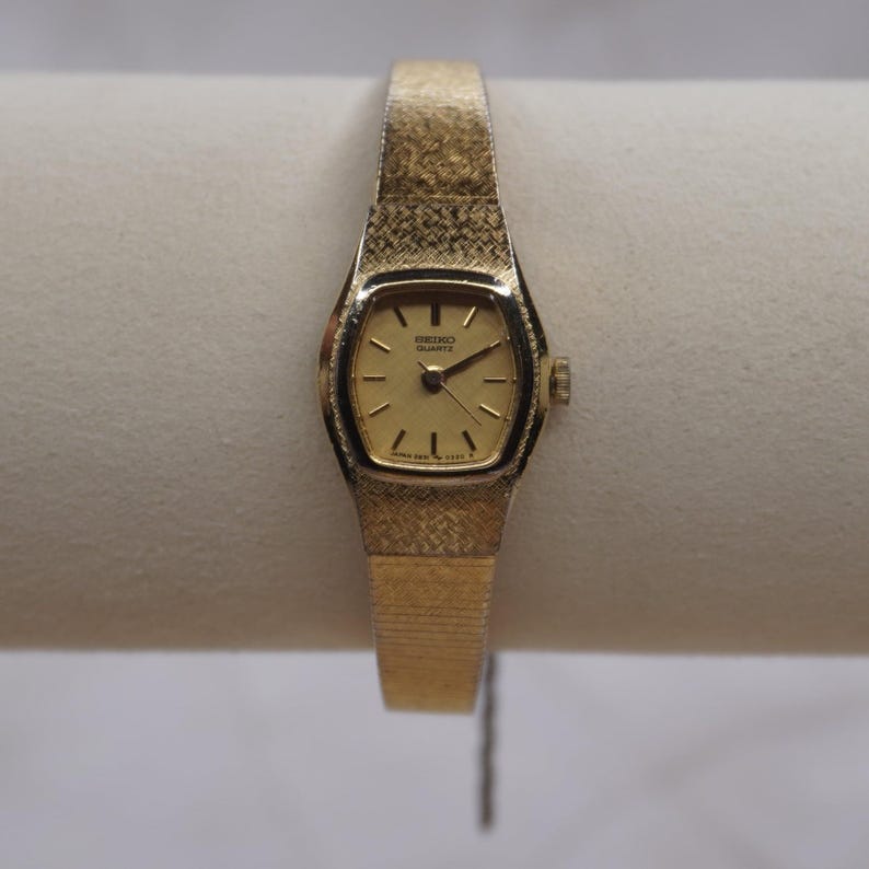 May include: A gold-toned Seiko Quartz watch with a rectangular face and a textured band. The watch face displays the Seiko Quartz logo and hands indicating the time. The band has a woven pattern.