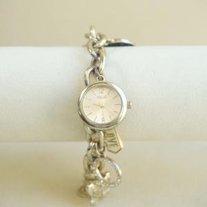 May include: A silver-tone Folio wristwatch featuring a round face with a light dial, thin hands, and numerical markers at twelve and six. The watch band is a chunky chain link style adorned with multiple dangling charms, including one with the word LOVE.