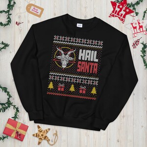 May include: Black sweatshirt with a festive, yet edgy design. The graphic features a goat head inside a yellow circle, with the words "HAIL SANTA" in white and red. The design includes Christmas trees and gift boxes, resembling a holiday sweater.