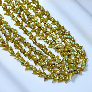 May include: A close-up shot of a necklace made of small, iridescent, golden-colored seashell beads. The beads are shaped like tiny shells and are strung together to form a necklace. The necklace is displayed on a white surface.