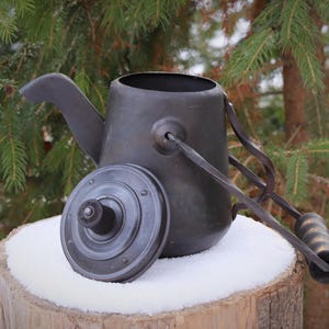 Könnte beinhalten: Eine dunkle Metallkanne mit Deckel, Griff und langem Griff mit Holzgriff. Die Kanne steht auf einem schneebedeckten Baumstumpf vor immergrünen Bäumen.