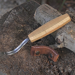 May include: A hook knife with a curved metal blade and a light brown wooden handle, accompanied by a small brown leather sheath featuring white stitching and two dark metal snap buttons for securing the blade when not in use.