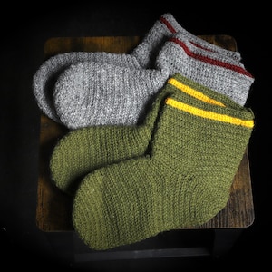 May include: Three pairs of knitted socks in shades of gray and green, arranged on a wooden surface. One pair is gray with a red stripe, another is green with a yellow stripe, and the third is a solid gray color. The socks are thick and appear warm.