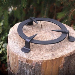 May include: A black, wrought iron trivet with a circular design and three supporting legs. The trivet is placed on a wooden tree stump, with a natural green background. The trivet is designed to hold hot pots and pans.