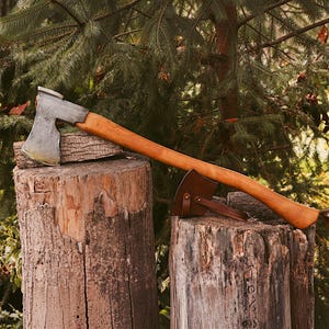 May include: A hand-forged steel axe head with a curved cutting edge attached to a long, smooth, light brown wooden handle. A dark brown leather sheath with a buckle strap is positioned near the base of the handle on a wooden log.