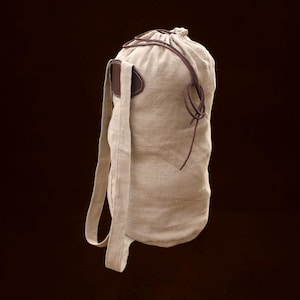 Puede incluir: Mochila de lino beige con un detalle de cuero marrón en el lateral y cierre de cordón de cuero marrón. La mochila tiene una larga correa beige para el hombro. La bolsa está hecha de una tela natural texturizada.