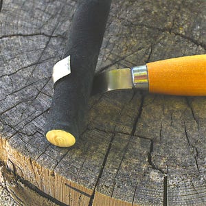 Könnte beinhalten: Ein Hakenmesser mit einer gebogenen Metallklinge und einem glatten, hellen Holzgriff. Die Klinge ist gegen ein schwarzes, zylindrisches Objekt positioniert, das am unteren Ende des Schafts ein kreisförmiges, helles Holzende aufweist.