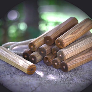 May include: A collection of cylindrical wooden dowels with a natural brown finish, each featuring a small centered hole at the end. The dowels are arranged in a stacked formation, with one separate piece lying horizontally and a thick rope visible.