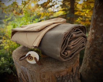 Può includere: Una coperta verde oliva arrotolata con una copertura beige è esposta su un ceppo d'albero. Un oggetto circolare bianco con un cinturino in pelle marrone e un logo è attaccato alla coperta. Lo sfondo presenta una vista sfocata di alberi e fogliame.