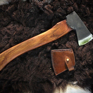 May include: A handcrafted axe with a polished steel blade and a wooden handle. The handle has a rich, warm brown tone. A brown leather sheath with a brass stud is included. The axe is displayed on a dark, textured surface.