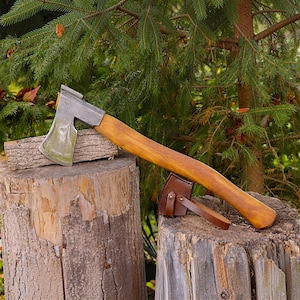 May include: A hand axe with a polished metal head and a smooth, curved wooden handle. A brown leather sheath with metal studs is attached to the handle near the head. The axe rests on a rough, cylindrical wooden log surface.