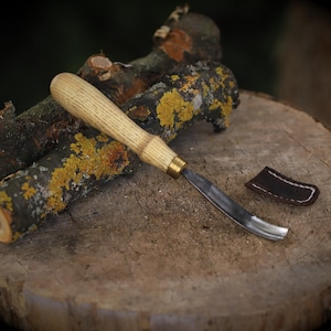 May include: A wood carving gouge with a curved blade and a light-colored wooden handle rests on a tree stump. A small, dark brown leather finger guard sits nearby. The gouge is a woodworking tool, used for shaping wood.