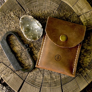 Pode incluir: Uma bolsa de couro castanho com fecho de pressão em latão e um pequeno logótipo de árvore estampado, acompanhada por uma pedra de sílex cinzenta e um percutor de aço forjado preto, dispostos sobre uma fatia circular de tronco.