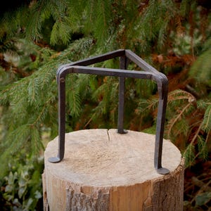 Può includere: Un portapiante artigianale in metallo scuro con una parte superiore triangolare e tre gambe curve. Il supporto è posto su un ceppo d'albero, con uno sfondo di rami di pino verdi.