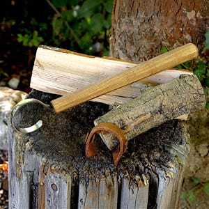 May include: A wood-handled hook tool with a polished metal curved blade, accompanied by a small brown leather strap, resting on a stack of split firewood logs. The tool features a long, cylindrical wooden handle and a smooth, reflective metal hook.