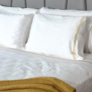 May include: White bed linen with a gold trim, a white pillow with a gold trim, and a brown knitted throw blanket.