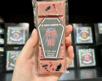 May include: A pink wax melt snap bar in clear plastic packaging, featuring embedded black bat shapes and red glitter. A coffin-shaped label reads Twisted Curiosities, Bite Me, and 1.7 oz | 48 g. The bar is divided into five rectangular segments.