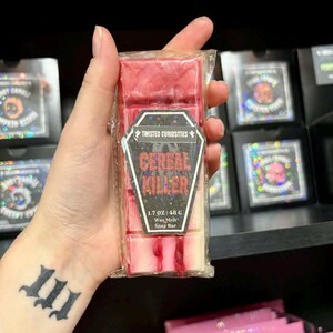 Puede incluir: Una barra de cera aromática roja y blanca en un paquete de plástico transparente. La etiqueta dice "Cereal Killer" con la marca "Twisted Curiosities". La barra pesa 48 g. Otros productos de cera aromática son visibles en el fondo.