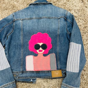 Può includere: Giacca di jeans blu con una grafica colorata sul retro. La grafica presenta una donna con afro rosa, occhiali da sole e una maglietta a righe rosse e bianche. Le maniche hanno toppe di tessuto a righe.
