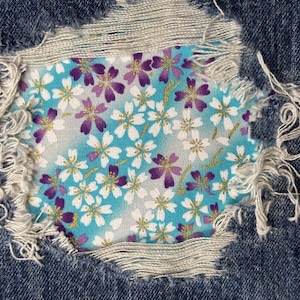 May include: A close-up of a ripped denim fabric with a floral pattern patch. The patch features white and purple flowers on a light blue background.