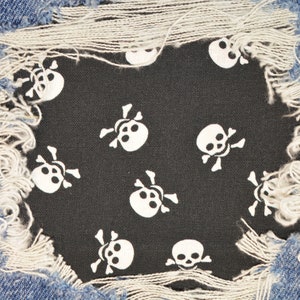 Puede incluir: Primer plano de un par de pantalones vaqueros azules desgarrados con un parche de tela negra debajo. El parche está cubierto con un patrón repetido de calaveras y huesos cruzados blancos.