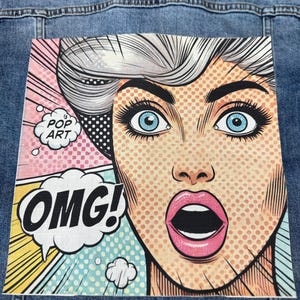 Könnte beinhalten: Jeansjacke mit Pop-Art-Grafik. Die Grafik zeigt eine überraschte Frau mit blauen Augen und dem Wort "OMG!" in einer Sprechblase. Die Worte "POP ART" sind ebenfalls in einer Sprechblase.