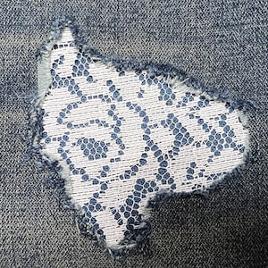 May include: A close-up of a pair of blue denim jeans with a white lace patch sewn over a hole in the fabric.