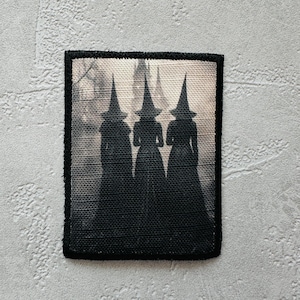 Può includere: Una toppa rettangolare nera con un'immagine color seppia di tre figure silhouette con cappelli a punta e lunghe vesti. Le figure sono in fila contro uno sfondo sfocato, creando un'estetica misteriosa e gotica. La toppa ha un bordo nero.