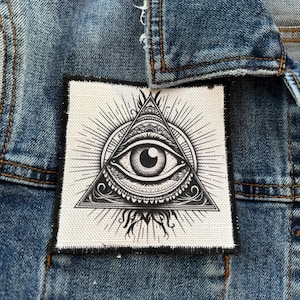 May include: A square, white patch with a black, detailed illustration of the all-seeing eye within a triangle, surrounded by sun rays. The patch is sewn onto a blue denim jacket.
