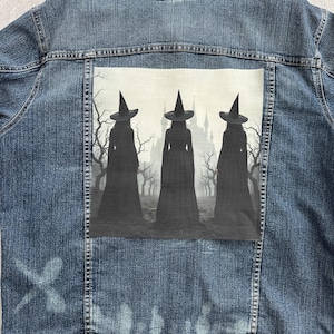 May include: A blue denim jacket with a graphic on the back depicting three silhouetted witches in front of a castle. The artwork is in shades of grey and black, creating a spooky Halloween theme.