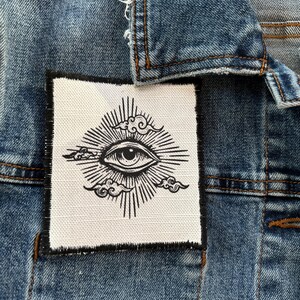 Può includere: Giacca di jeans blu con una toppa bianca con un disegno di occhio nero, circondato da raggi di sole e nuvole. La toppa è cucita sul tessuto blu della giacca, mostrando un dettaglio artistico unico. La giacca ha cuciture classiche.
