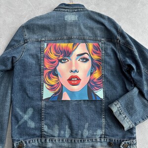 Może przedstawiać: Jeansowa kurtka w kolorze niebieskim z portretem w stylu pop-art na plecach. Portret przedstawia kobietę z czerwonymi ustami, niebieskimi oczami i kolorowymi włosami. Kurtka jest w średnim odcieniu niebieskiego z guzikami i kieszeniami.