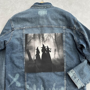 Könnte beinhalten: Hellblaue Jeansjacke mit einem schwarz-weißen Aufdruck von drei Hexen in einer Waldszene auf dem Rücken. Die Jacke hat einen Knopfverschluss, einen klassischen Kragen und zwei Brusttaschen. Die Jacke hängt an einem Holzbügel.