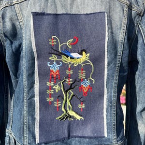 Puede incluir: Una chaqueta vaquera con un parche bordado azul que representa un pájaro posado en una rama con flores rojas y verdes.