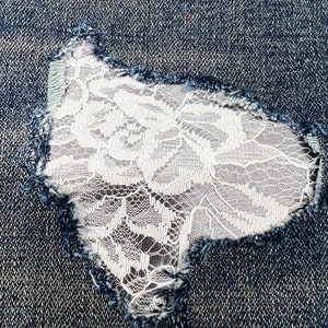 May include: Close-up of distressed denim fabric with a tear patched with white floral lace. The edges of the tear are frayed, and the lace is visible through the opening, creating a contrast in texture and color.