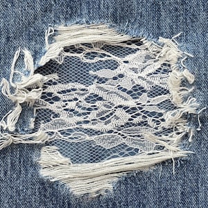 Può includere: Un primo piano di un tessuto denim strappato con una toppa di pizzo bianco cucita sopra il buco.