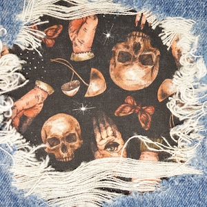 Peut inclure: Gros plan sur un tissu en jean déchiré avec un patch de tissu noir en dessous. Le patch présente un motif répété de crânes, de mains et de papillons dans des tons d'or, de brun et de blanc.