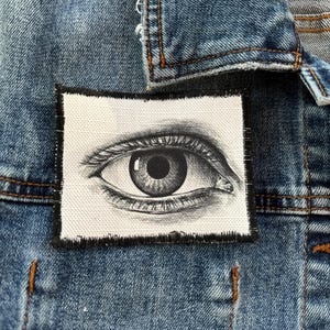 May include: A denim jacket with a rectangular patch showing a detailed black and white illustration of an eye. The patch is sewn onto the jacket, which has visible stitching and a collar.