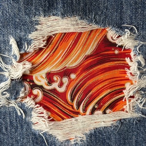 Puede incluir: Primer plano de un par de pantalones vaqueros azules rasgados con un parche de tela colorido debajo. El parche de tela presenta un patrón de ondas rojo, naranja y dorado.