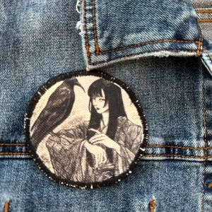 May include: A round patch featuring a black and white illustration of a woman in traditional clothing with a raven. The patch is sewn onto a blue denim jacket.