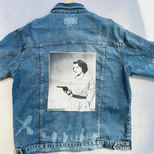 May include: A light blue denim jacket featuring a black and white image of a woman holding a gun. The jacket has a classic design with a button-up front, pockets, and a collar. The image is printed on the back of the jacket.