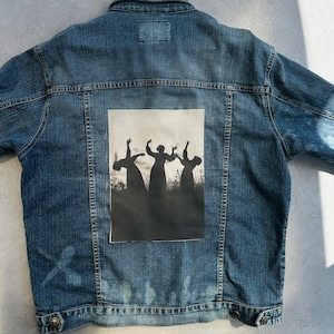 Op de afbeelding: Een blauw spijkerjack met een zwart-wit afbeelding van drie dansende figuren op de rug. De jas heeft een klassiek ontwerp met een knoopsluiting, zakken en een kraag. De denim is een medium wassing.