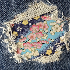 May include: A close-up of a denim fabric with a floral pattern patch sewn on. The patch is made of a light blue fabric with pink and white flowers. The denim fabric is ripped and frayed around the edges of the patch.