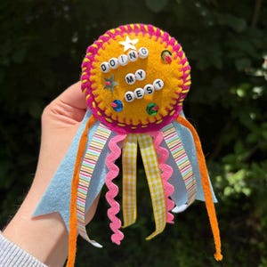 Puede incluir: Una escarapela circular de fieltro en naranja brillante con un borde festoneado rosa intenso. El centro presenta cuentas de letras blancas que deletrean DOING MY BEST, adornadas con pequeñas estrellas y lentejuelas. Cuelgan cintas azul claro, naranja y amarillas.