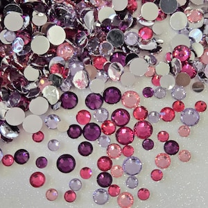 May include: Assortment of pink, purple, and clear rhinestones. The rhinestones are scattered on a white surface.