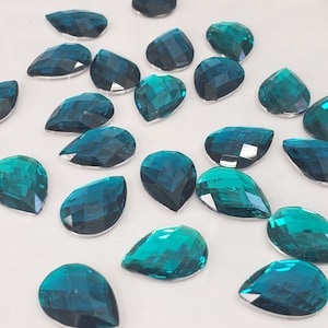 May include: A collection of teal teardrop-shaped rhinestones. The rhinestones have a faceted design and are scattered on a white surface.