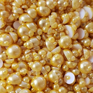 May include: A close-up of a variety of pearl-like beads in a golden color. The beads are of different sizes and shapes, some are round, some are half-round, and some are flat.