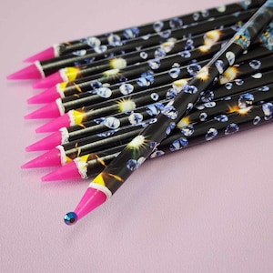 May include: A group of black pencils featuring a pattern of blue gems and yellow bursts. Each pencil has a vibrant pink tip and a small, iridescent bead. The pencils are displayed on a light pink surface.
