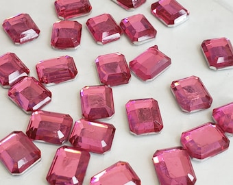 May include: A collection of pink, faceted, rectangular rhinestones. The rhinestones are scattered on a white surface.